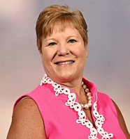 A woman wearing a pink shirt and a pearl necklace is smiling for the camera.