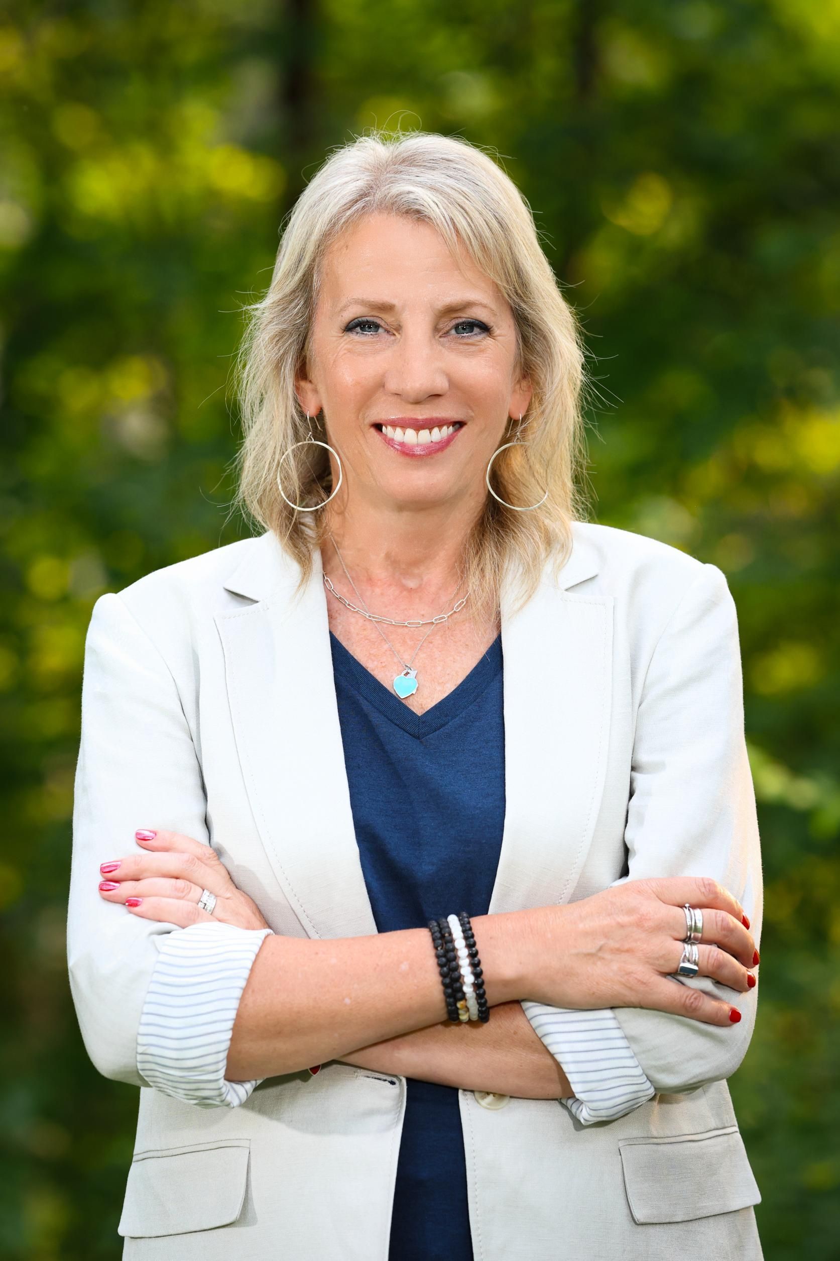 A woman in a white jacket and blue shirt is standing with her arms crossed and smiling.