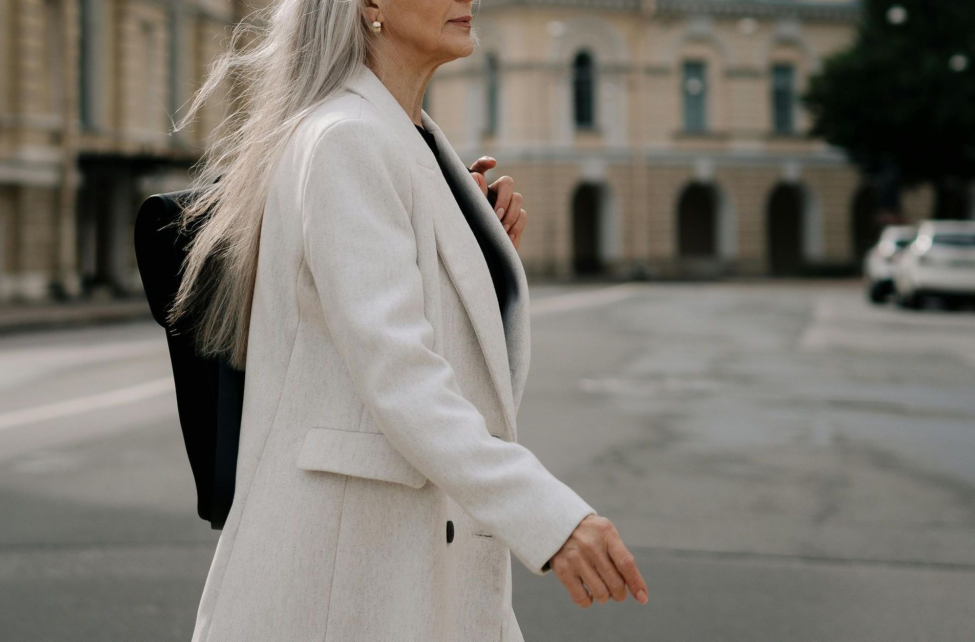A woman with gray hair is walking down the street wearing a white coat.