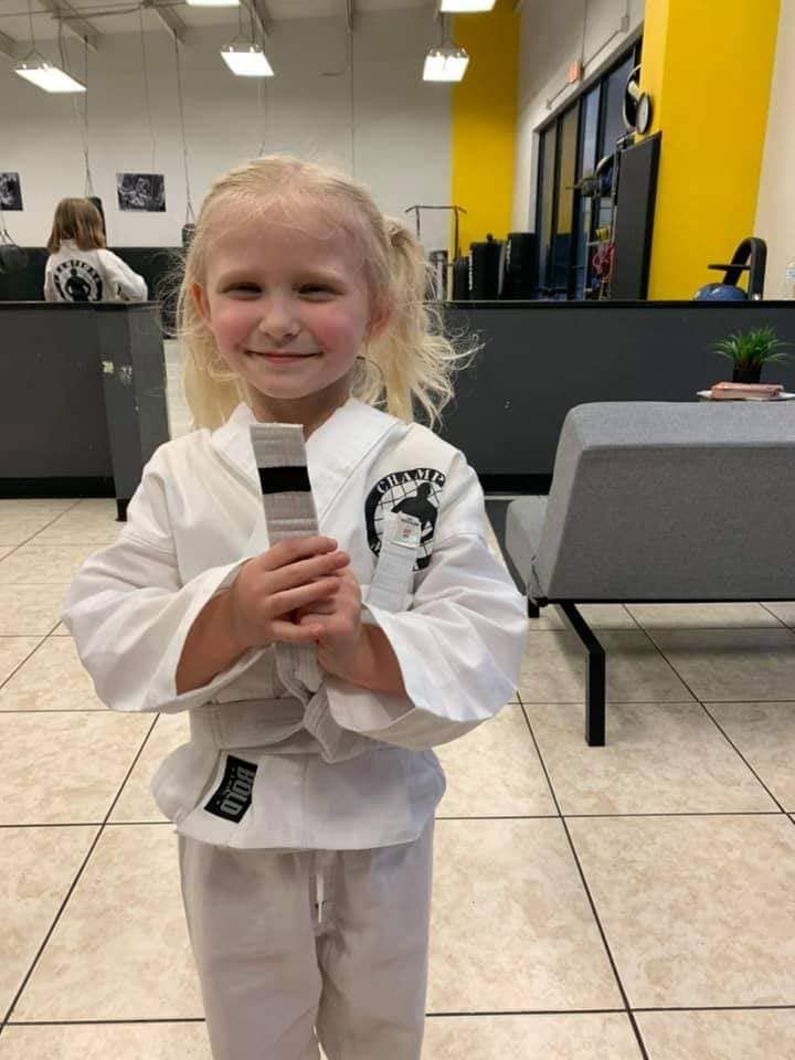 A little girl in a white karate uniform is holding a black belt.