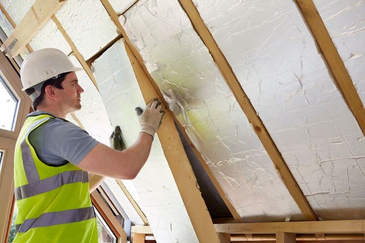 Worker in hard hat installing reflective insulation between roof rafters inside a house frame
