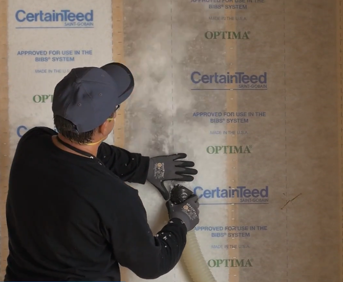 Worker in hard hat and blue shirt spraying insulation inside a wooden-framed house wall