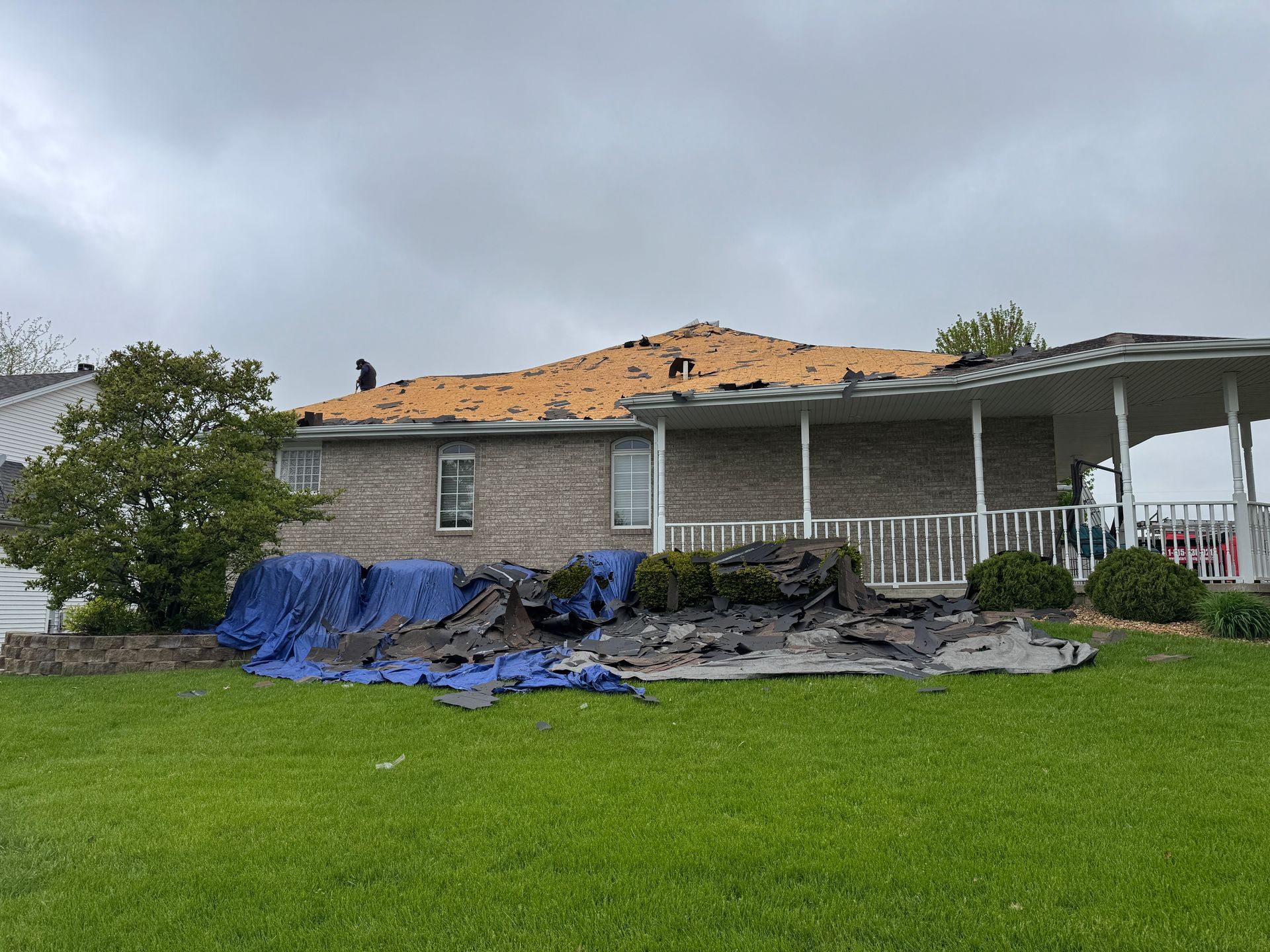 A brick house with a partially removed roof exposing the wooden deck, surrounded by blue tarps on the lawn.