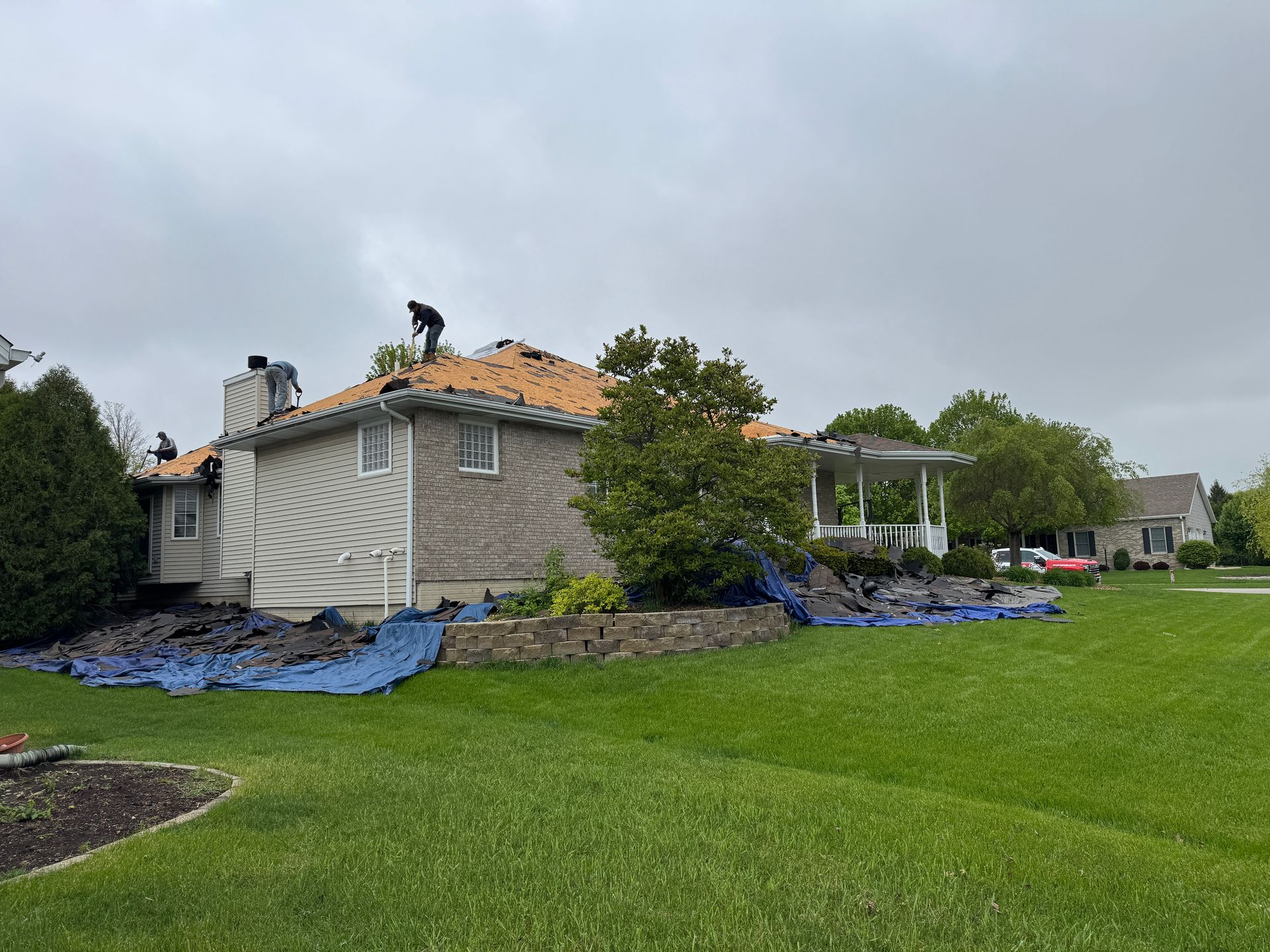 Construction workers replace the roof of a suburban house, with blue tarps catching debris on the lawn.