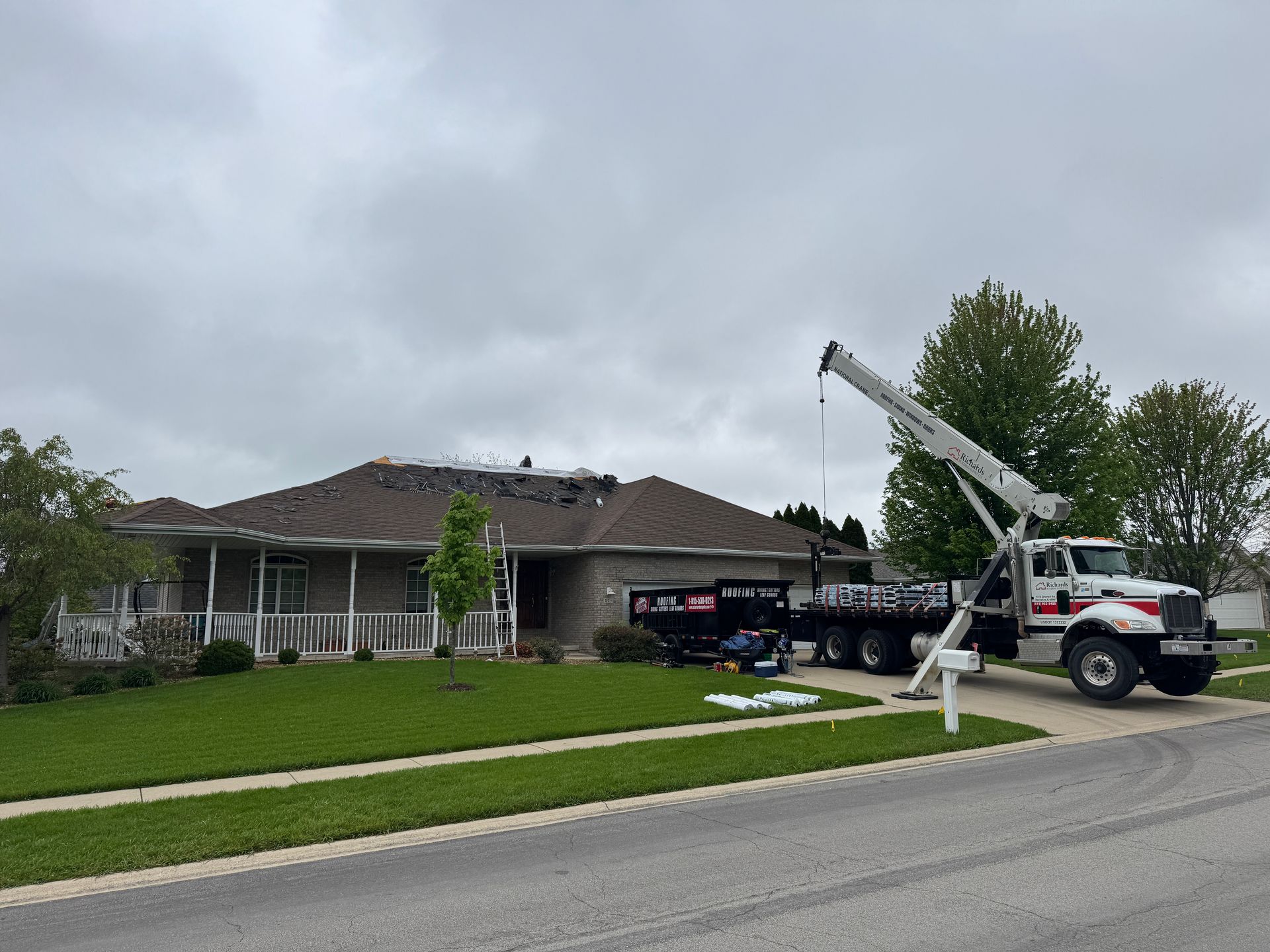 A white construction crane parked on a suburban street next to a house undergoing roof repairs, with a dumpster nearby.