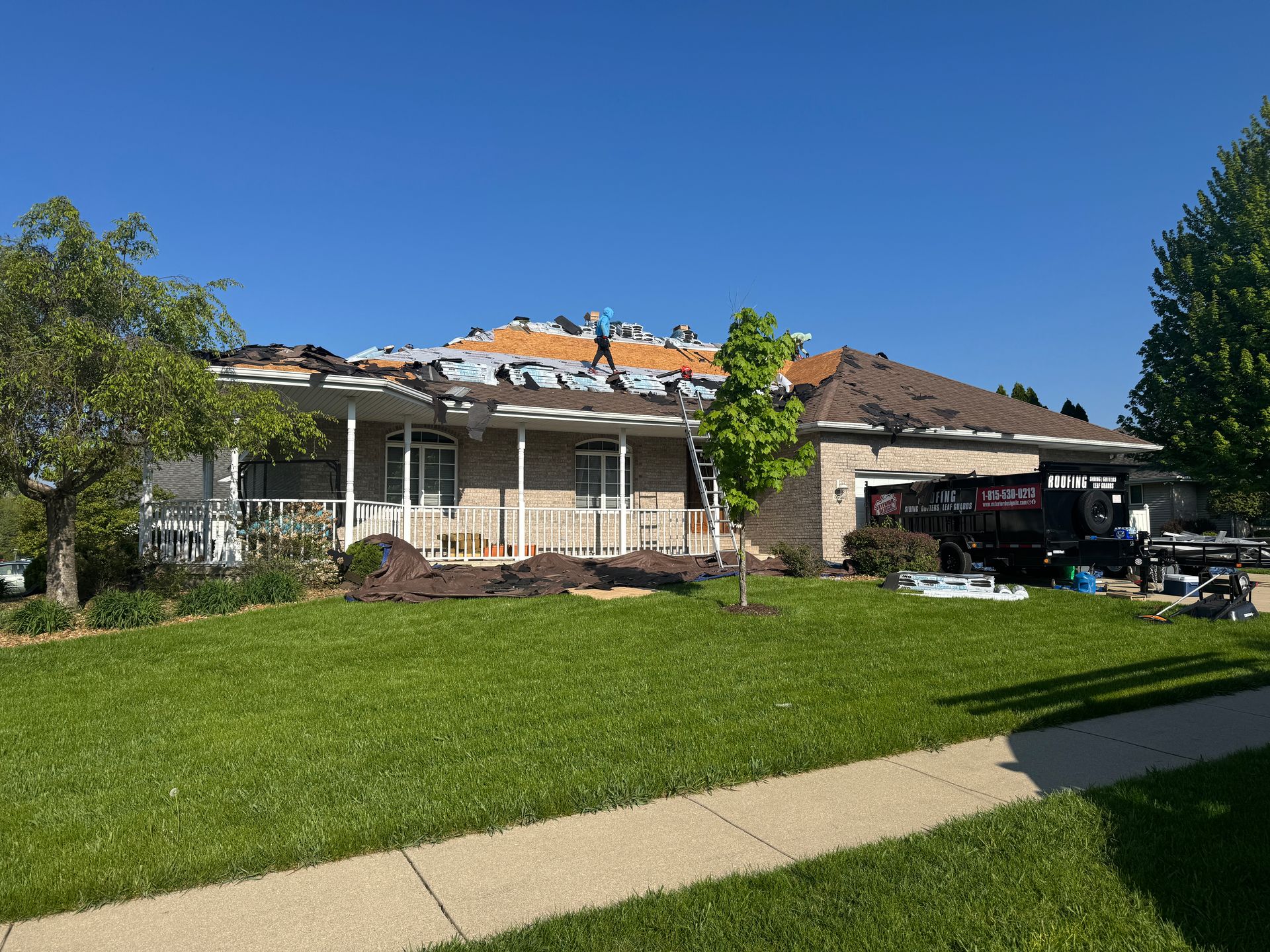 Workers replace shingles on a residential brick house roof under a clear blue sky, with a disposal dumpster parked nearby.
