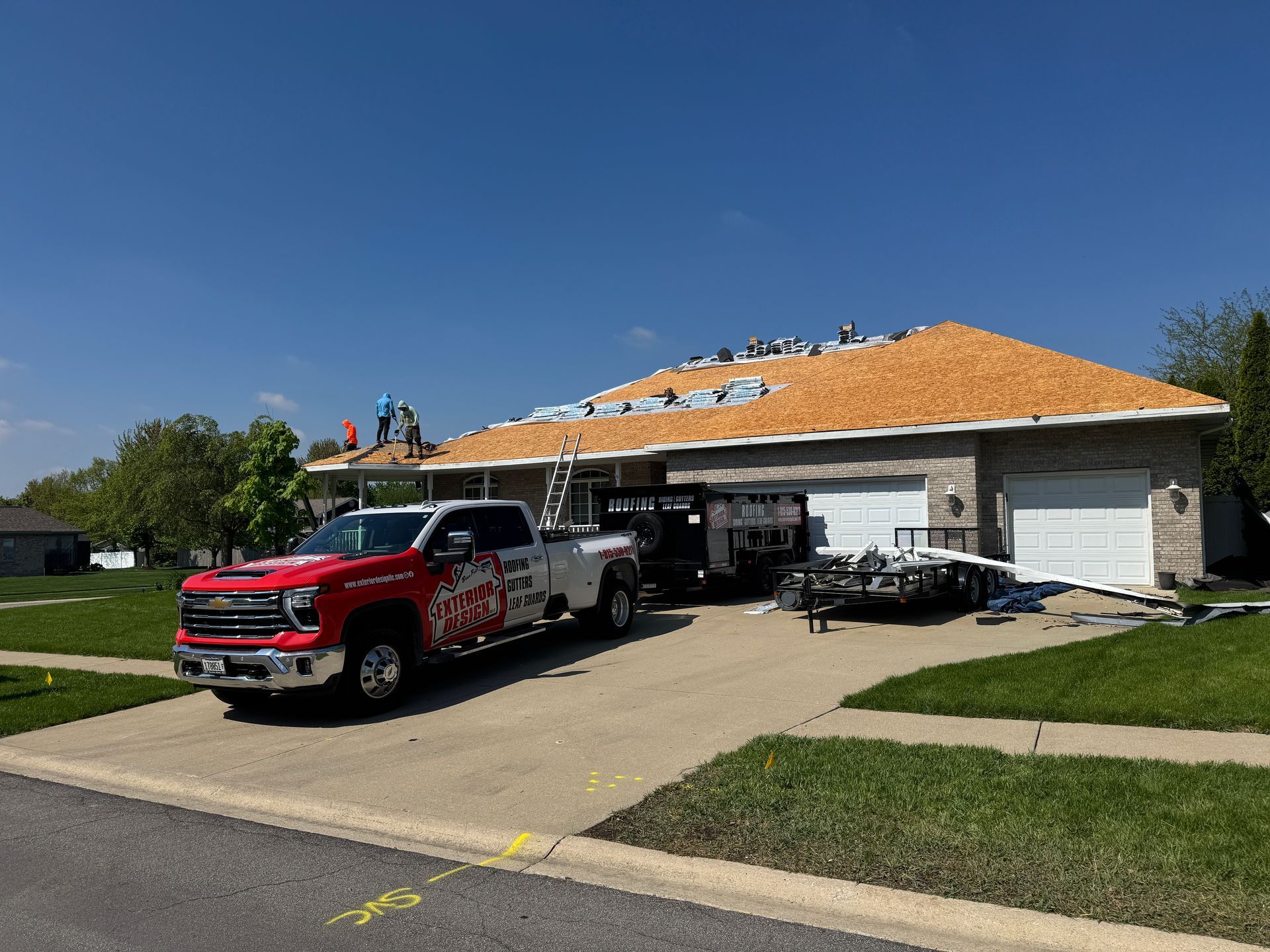 Workers on the roof of a suburban home undergoing repairs, with a red company truck and dumpster in the driveway.