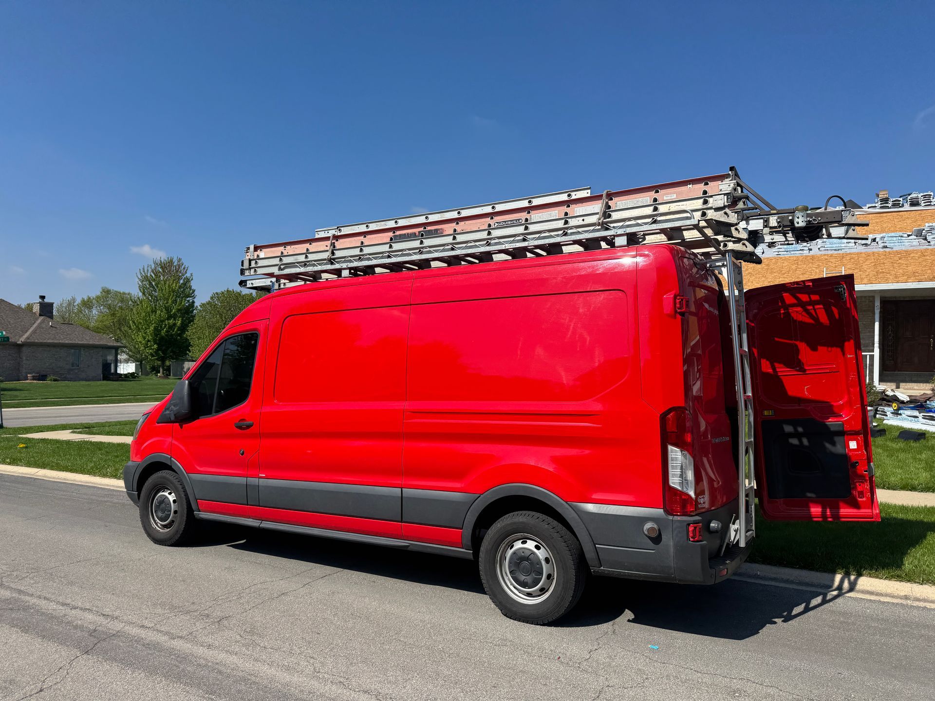 A bright red work van with a ladder rack parked on a suburban street, with its rear doors open.