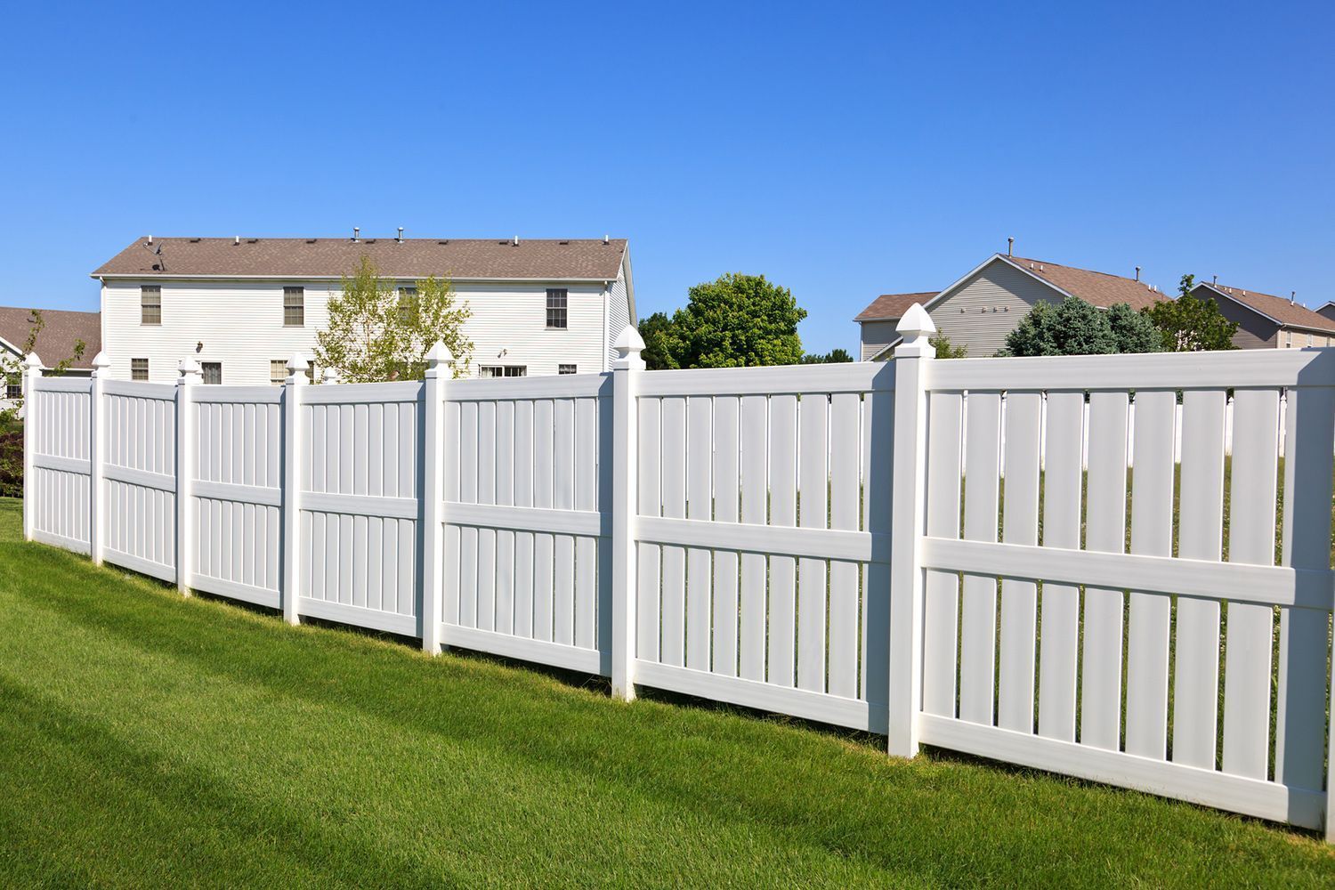 White wooden fence bordering a green lawn in a suburban neighborhood with houses in the background