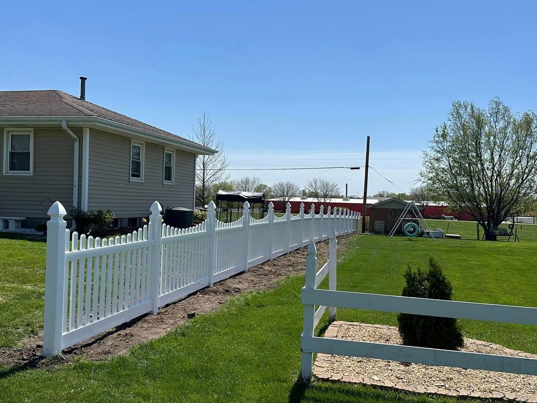 White fence along a grassy yard beside a beige house under a clear blue sky