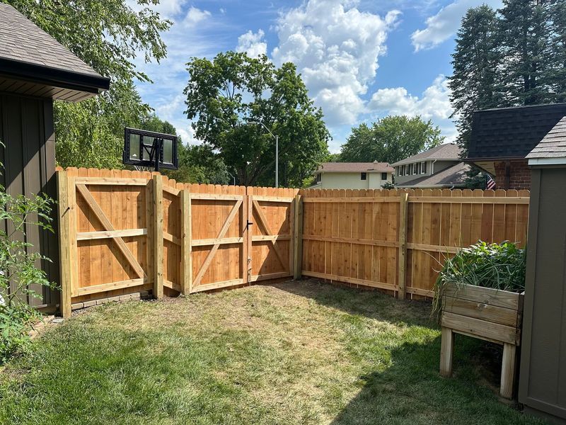 Backyard with wooden fence, open gate, and patchy grass under a partly cloudy sky