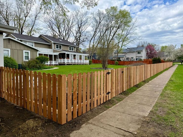 Wooden picket fence around a green yard beside a sidewalk, with houses and trees under a cloudy sky