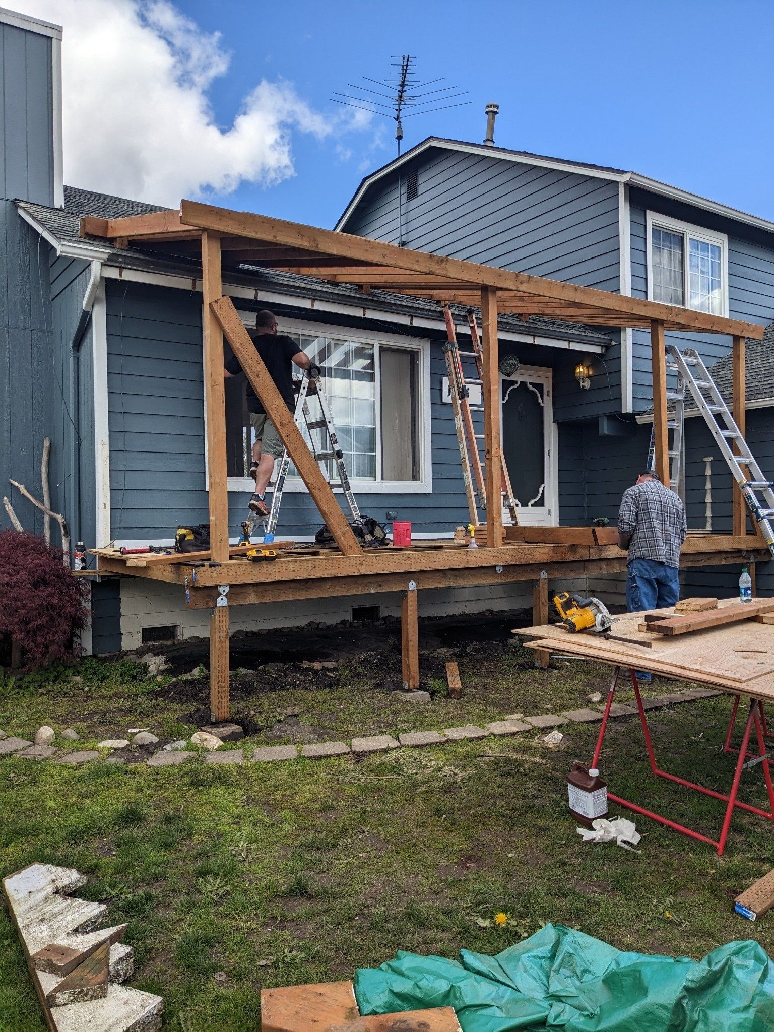 Partially built wooden porch on a blue house, with a worker standing nearby in a yard.