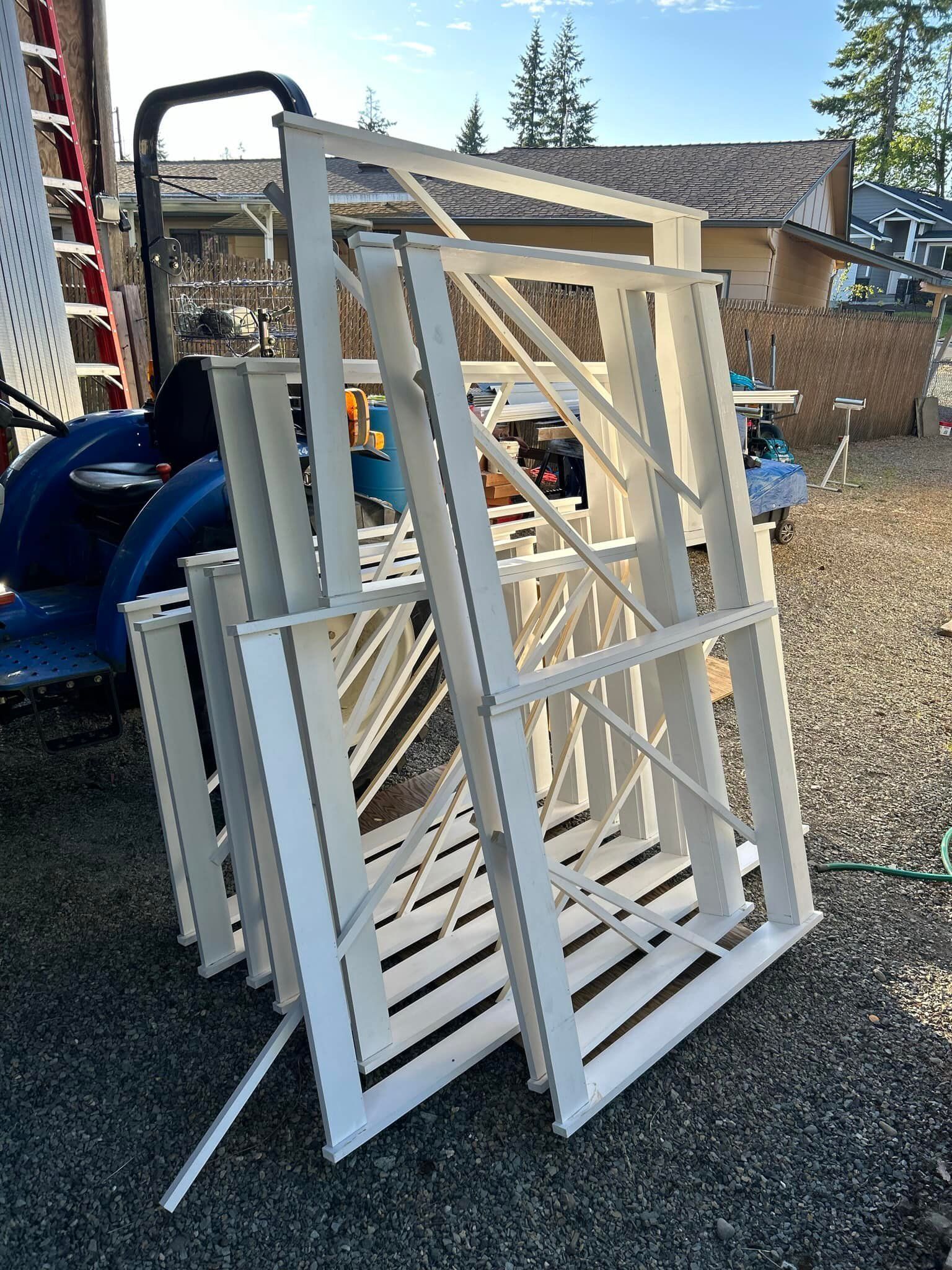 Stacked white window frames and screens leaning outdoors on gravel beside a house.