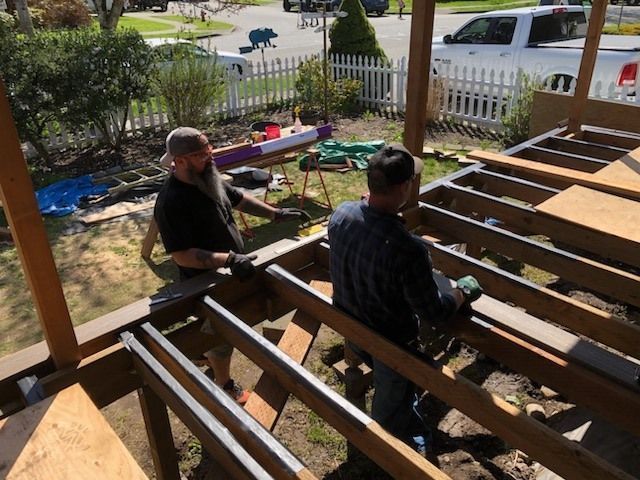 Two workers framing a wooden deck on a sunny porch with tools and materials nearby.