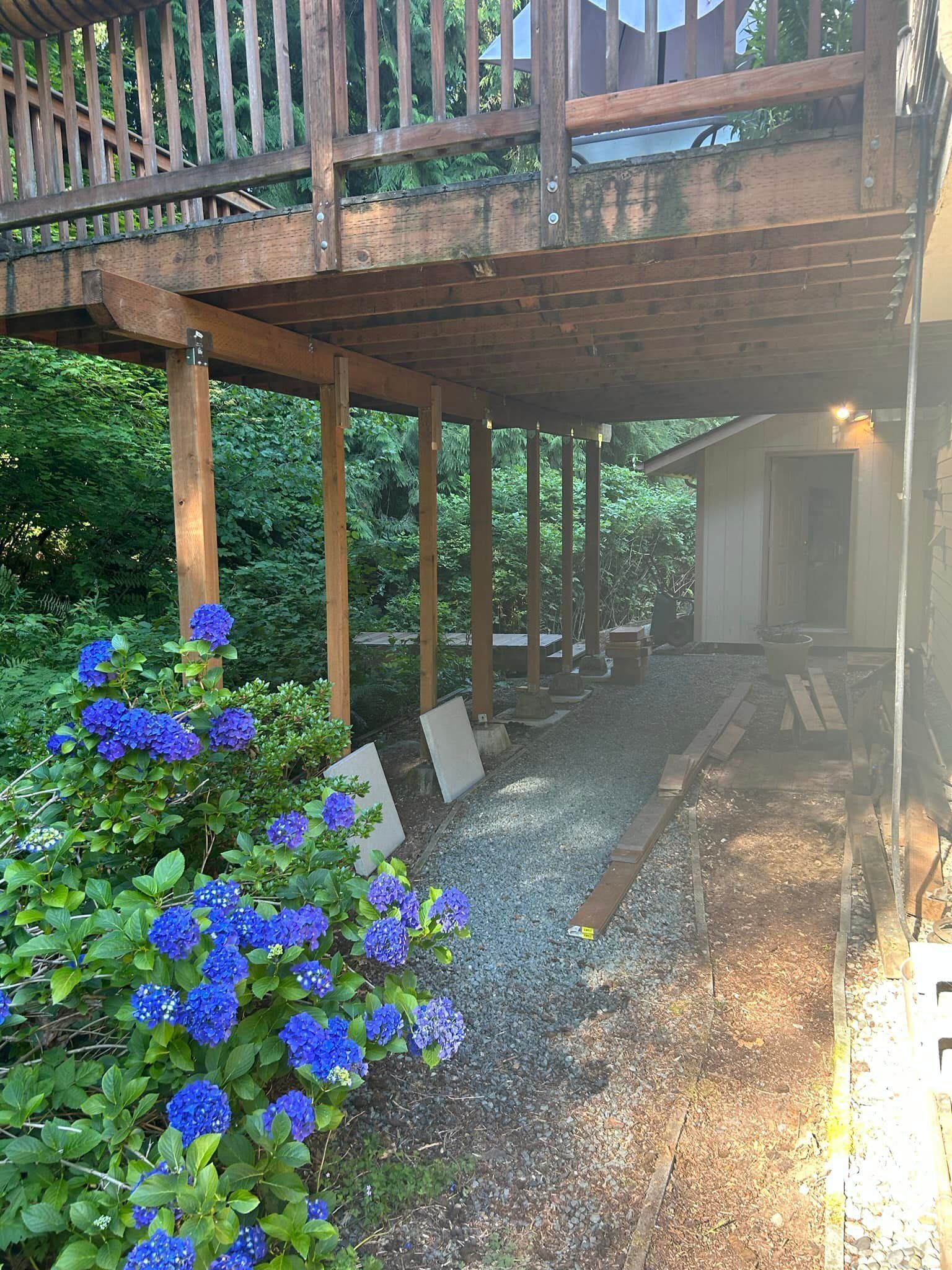 Wooden deck over gravel path beside a white shed, with blue flowers and sunlight filtering through trees.