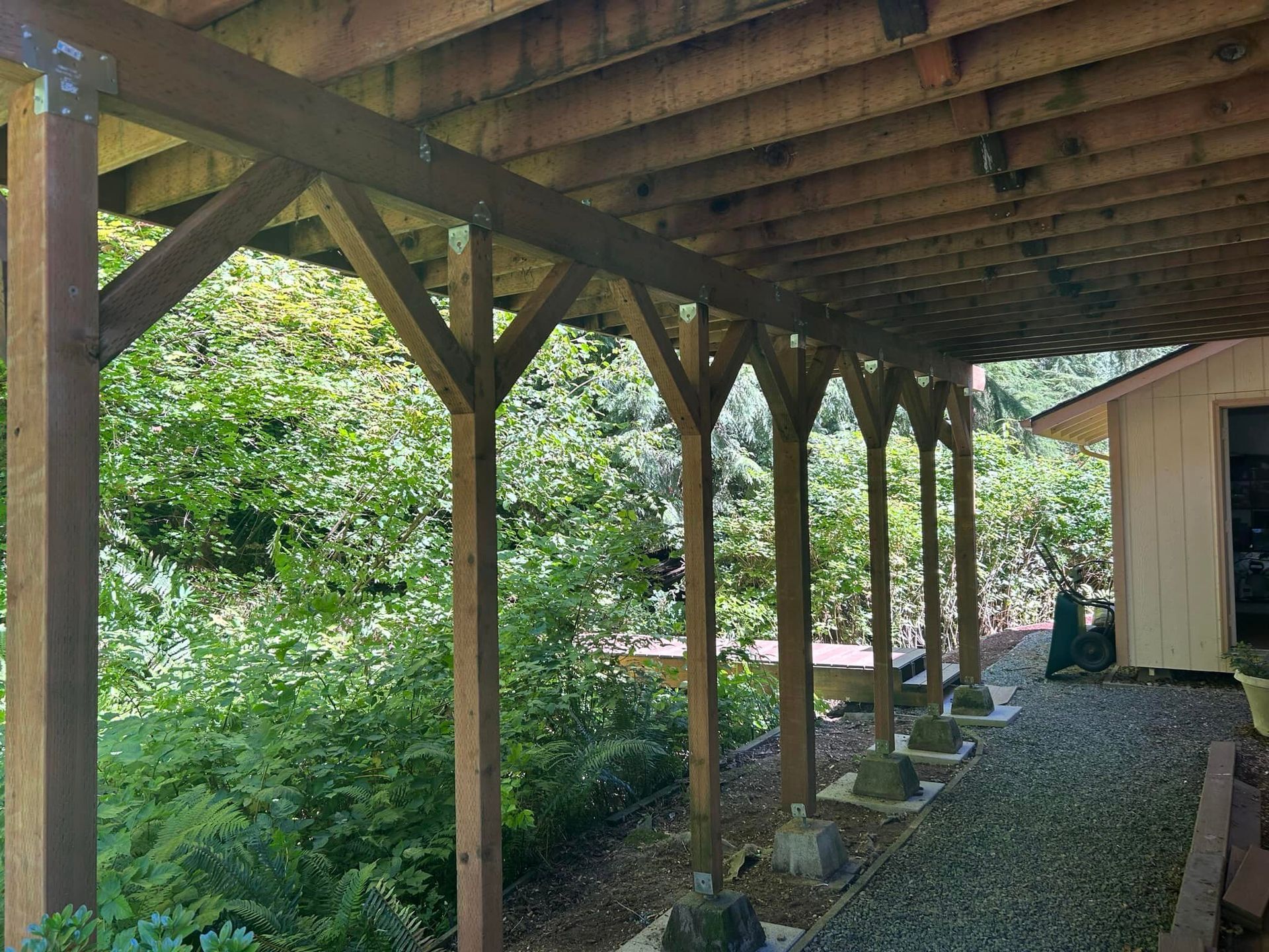 Wooden covered patio walkway with posts, gravel ground, and dense green foliage beside a shed