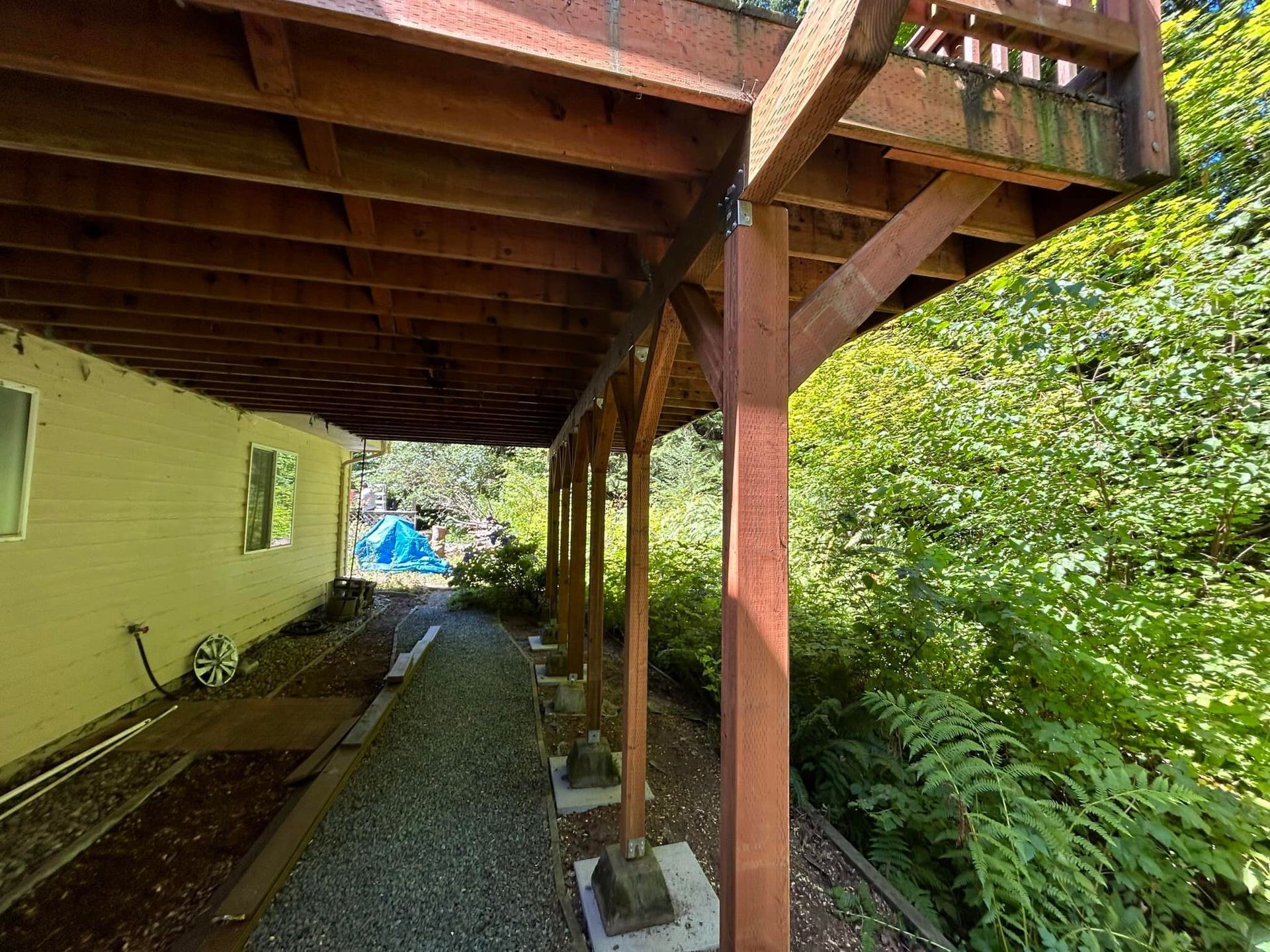 Wooden deck beside a yellow house, overlooking a gravel path and dense green foliage