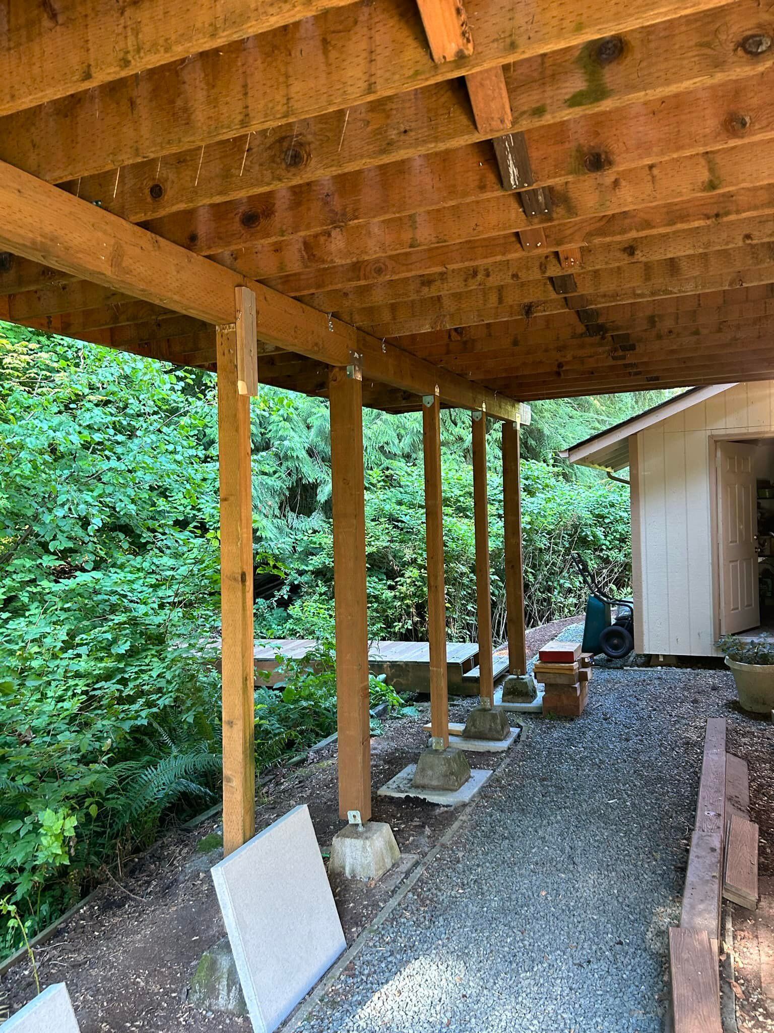 Covered gravel walkway beside a wooden deck, with support posts, greenery, and a small shed.