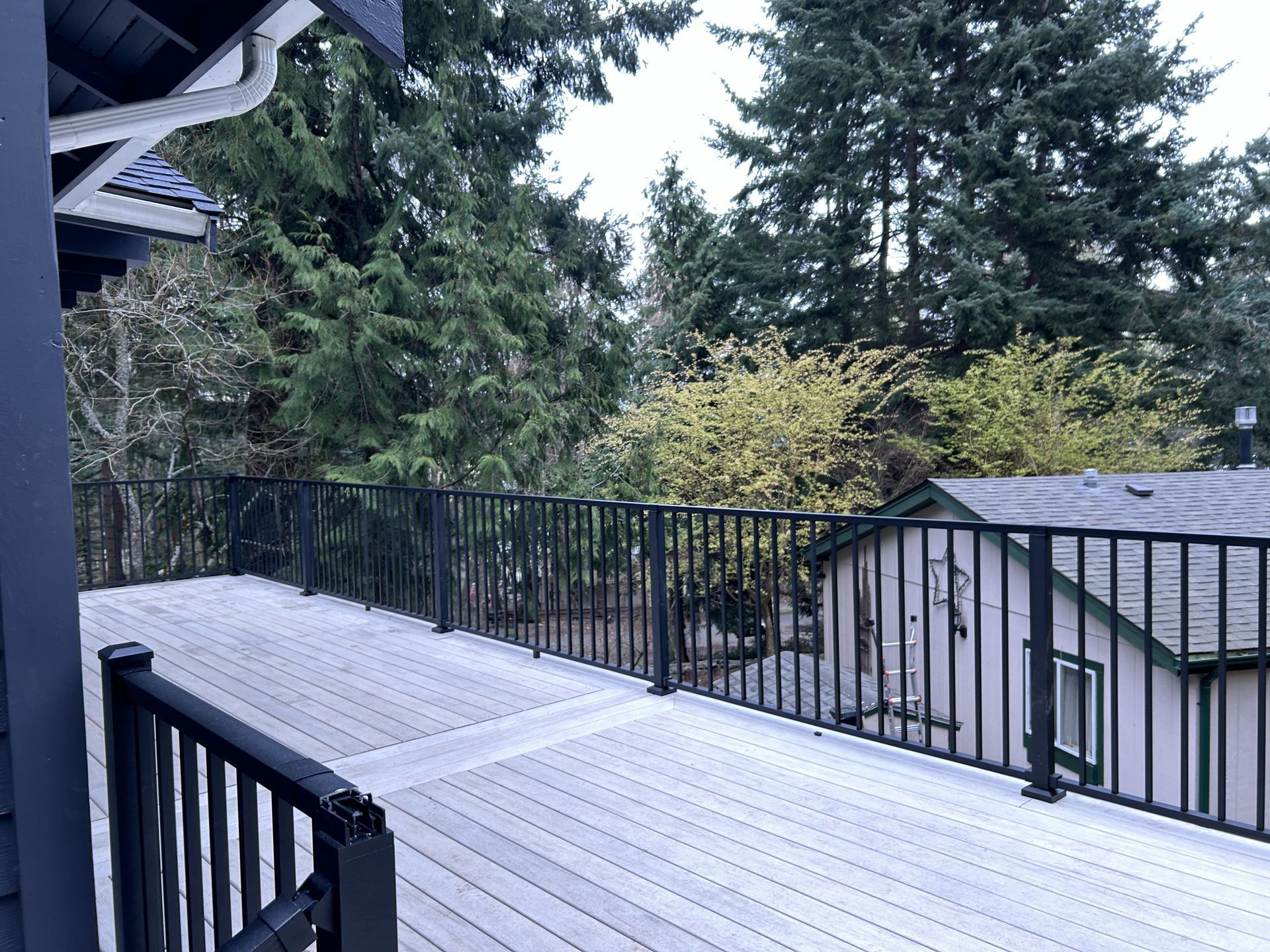 Wooden deck with black railing overlooking trees and a neighboring house on a cloudy day