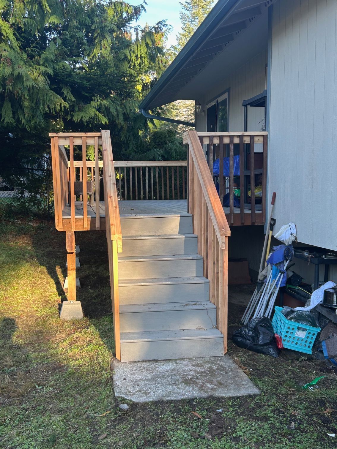 Concrete steps with wooden railings leading to a raised house porch, with yard and stored items beside it.