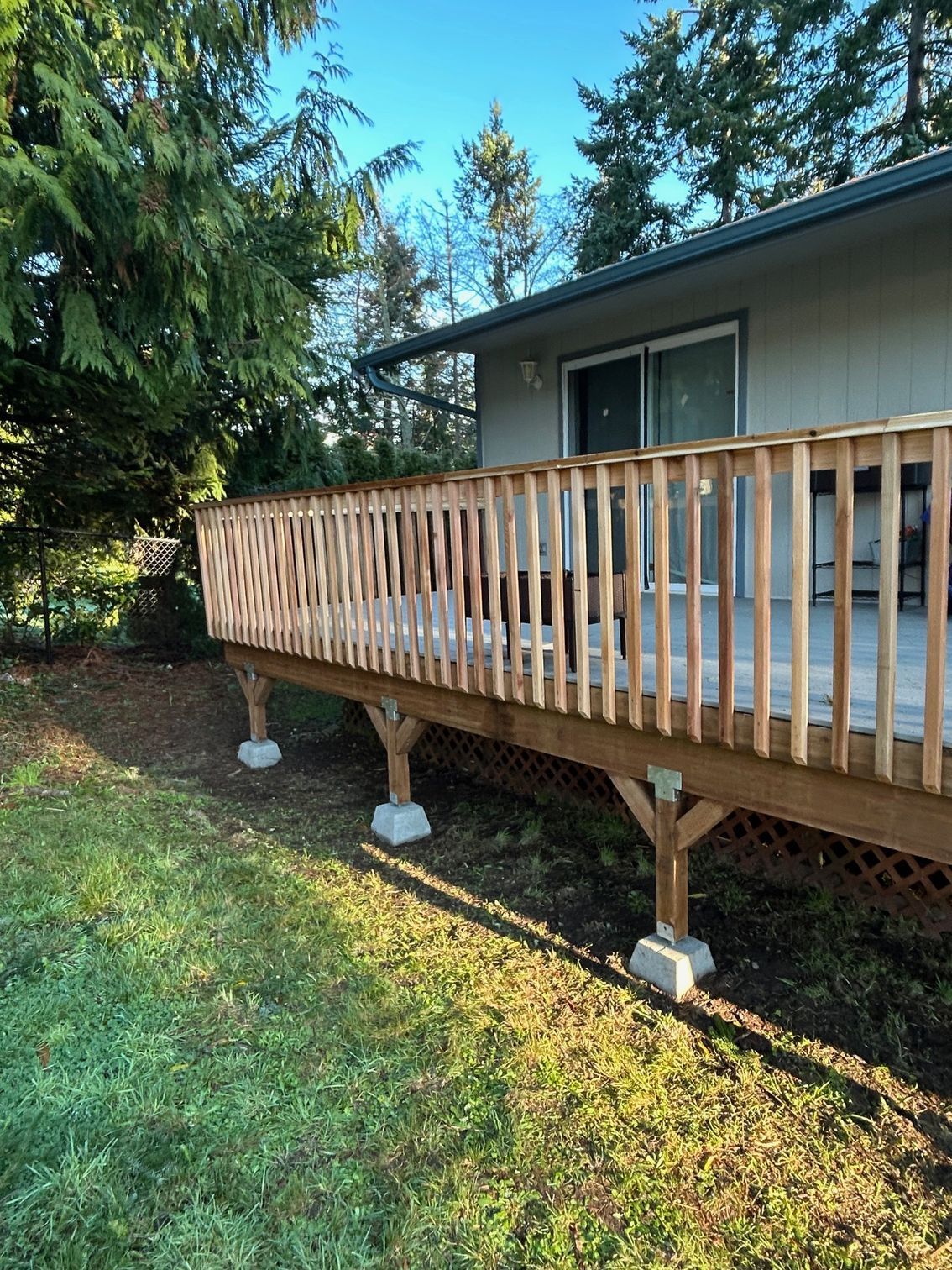 Raised wooden deck with railing attached to a house, supported by concrete footings in a grassy yard.