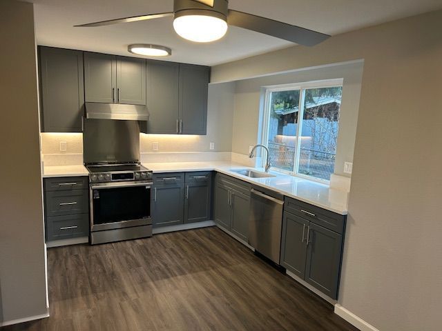 Modern kitchen with gray cabinets, stainless appliances, white counters, and a window over the sink.
