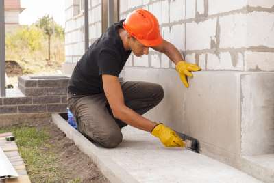 A worker applying a waterproof foundation sealer to the base of an exterior wall to avoid a foundation leak repair.