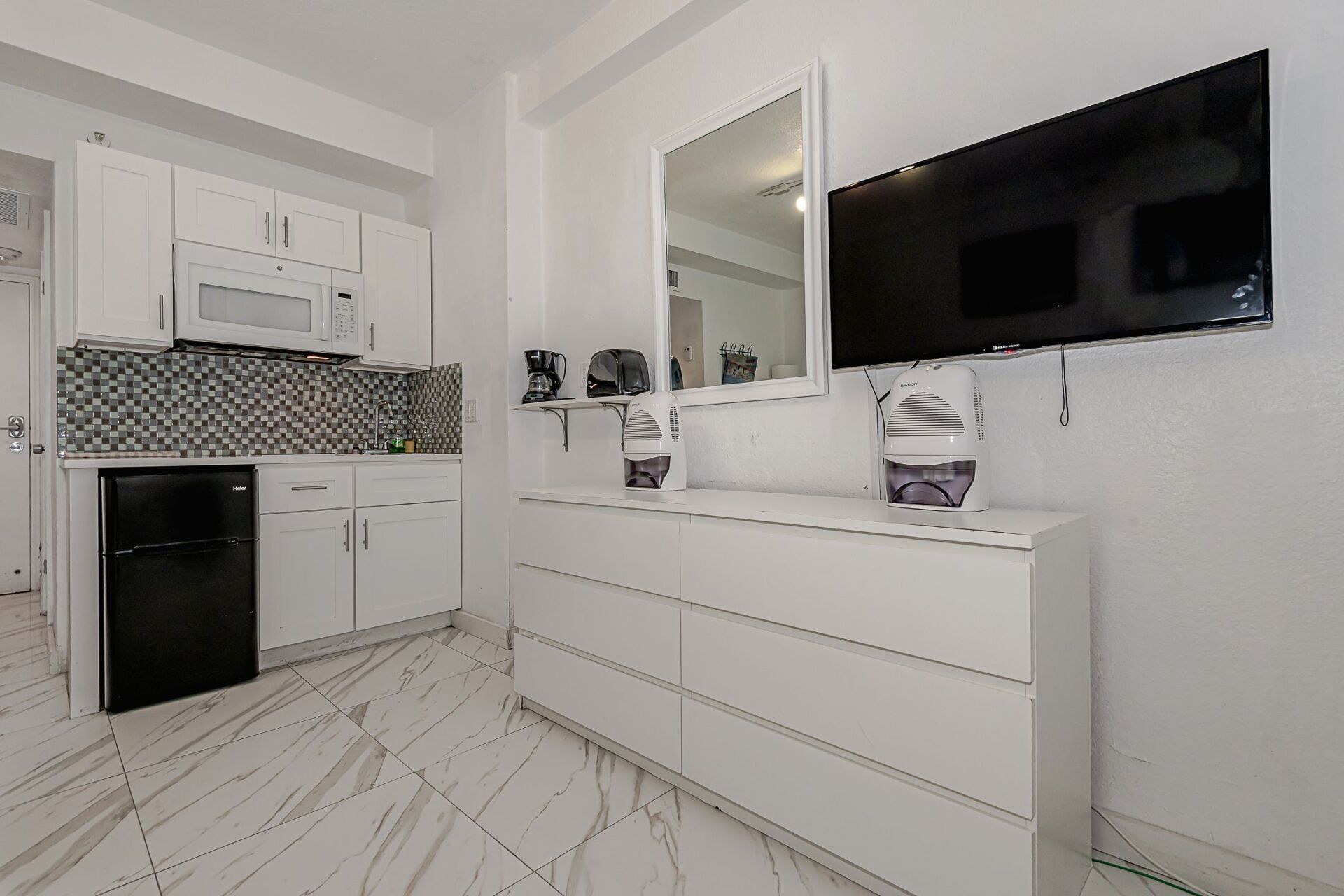 A kitchen with white cabinets , a black refrigerator , a flat screen tv , a mirror and a dresser.