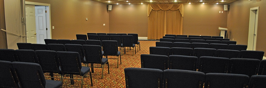 Rows of dark chairs in a room with a stage. Golden curtain on the stage; beige walls; tan carpet.