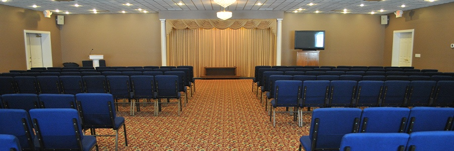 An empty auditorium with rows of blue chairs, tan walls, beige curtains, and a brown carpet.