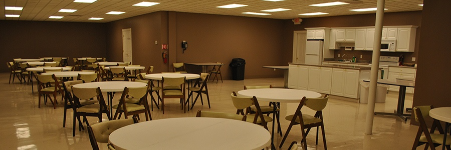 A large room with round tables and chairs, presumably a dining or meeting space. There is a kitchen in the background.