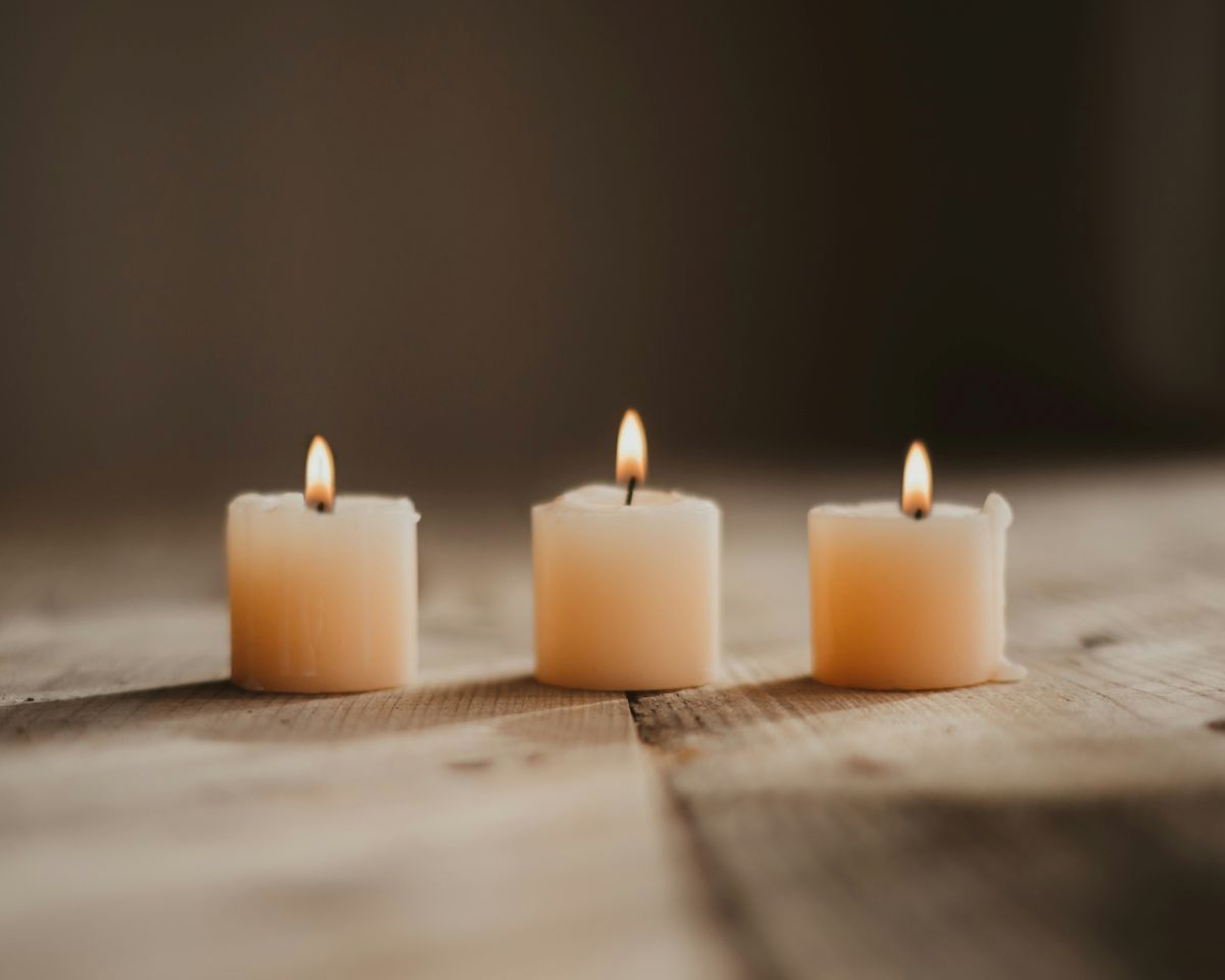 Three lit, square candles on a wooden surface, with a blurred, neutral background.