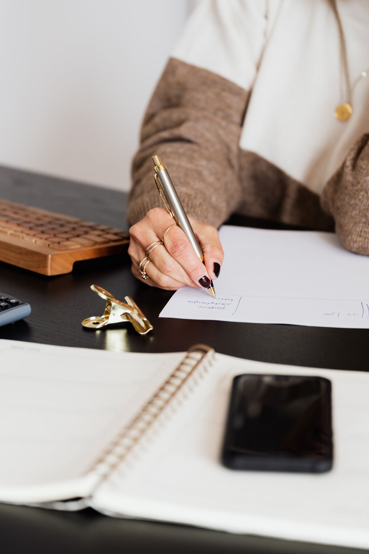 Person writing on paper at a desk with a phone, notebook, calculator and gold clip.