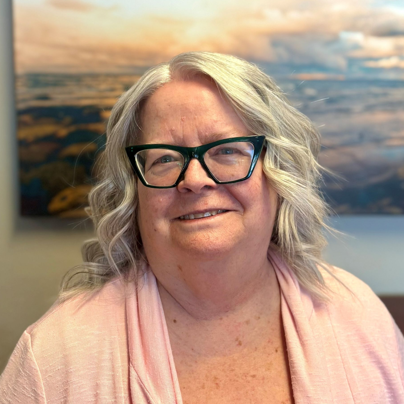 Portrait of a woman with short white hair and glasses, in a blue shirt and red scarf, in front of a blurry green background.