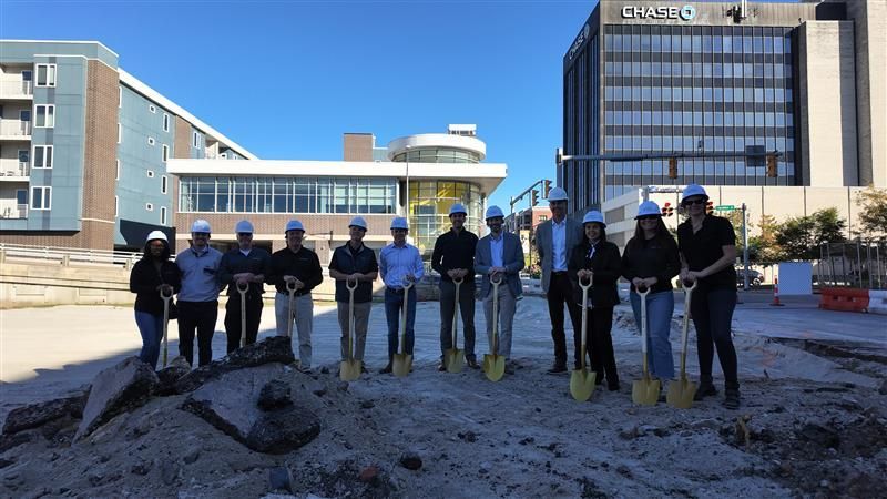 People with shovels at a groundbreaking ceremony, standing in front of buildings.