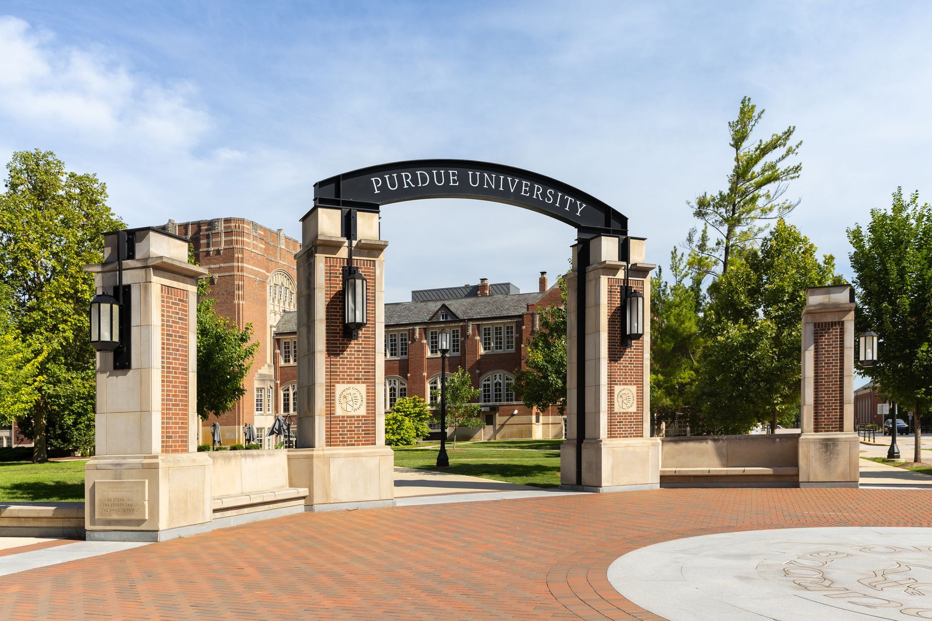 Purdue University entrance archway with brick pillars, red brick road, and buildings under blue sky.