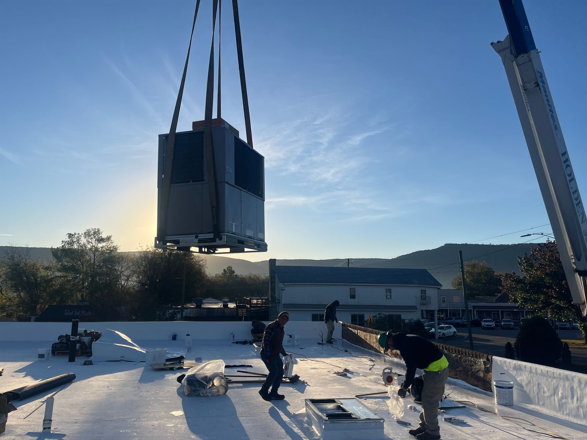 Construction crew installing a large rooftop HVAC unit with a crane at sunrise, workers preparing materials on a commercial building roof,