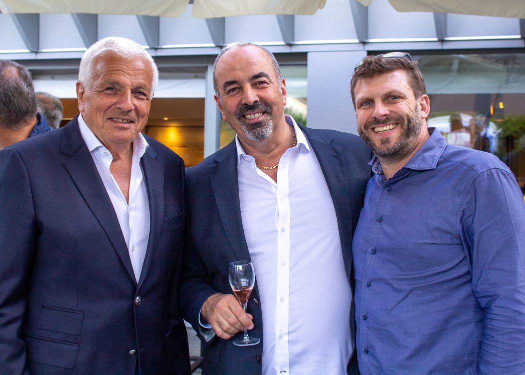 Trois hommes posent pour une photo ensemble et l'un d'eux tient un verre de vin.