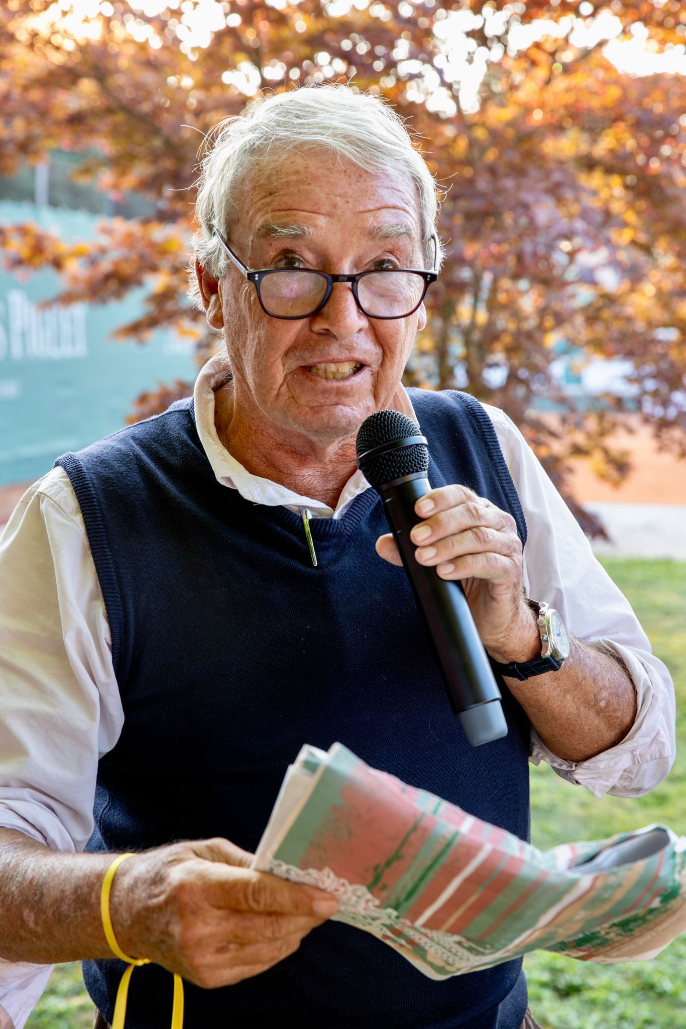 Un homme portant des lunettes tient un microphone et lit un journal.