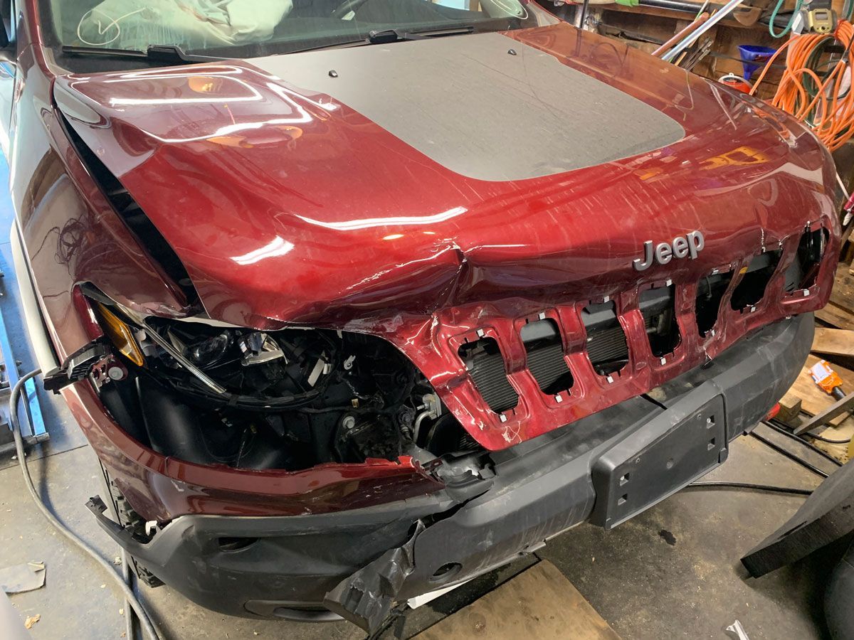 Damaged red Jeep front, crumpled hood, headlight broken, in a repair shop.