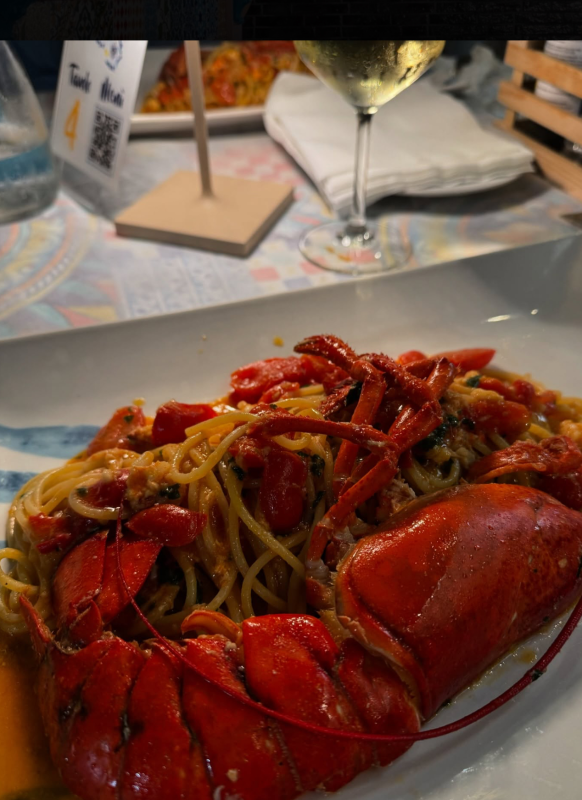 Piatto di pasta all'astice servito su un piatto bianco, con un bicchiere di vino bianco sullo sfondo.