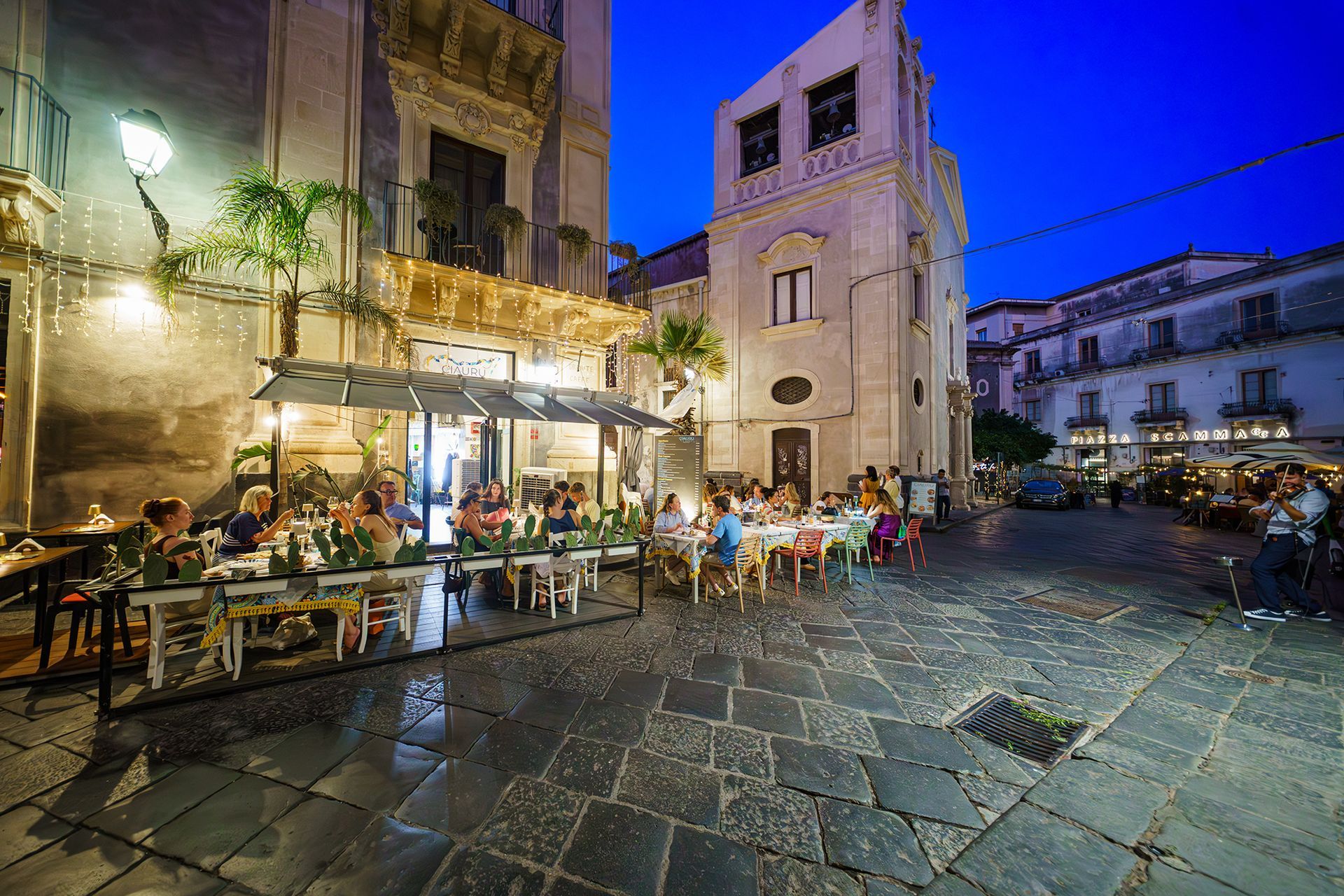 Ristorante all'aperto al tramonto in una piazza di una città europea, con commensali, tavoli ed edifici.