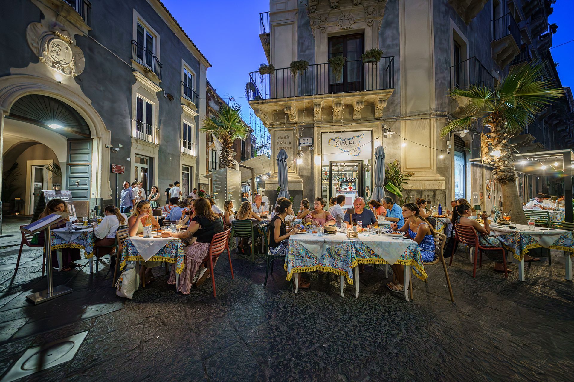 Tavoli di un ristorante su una strada acciottolata al tramonto, con persone che cenano all'aperto. Tovaglie blu e bianche, illuminate da lucine.