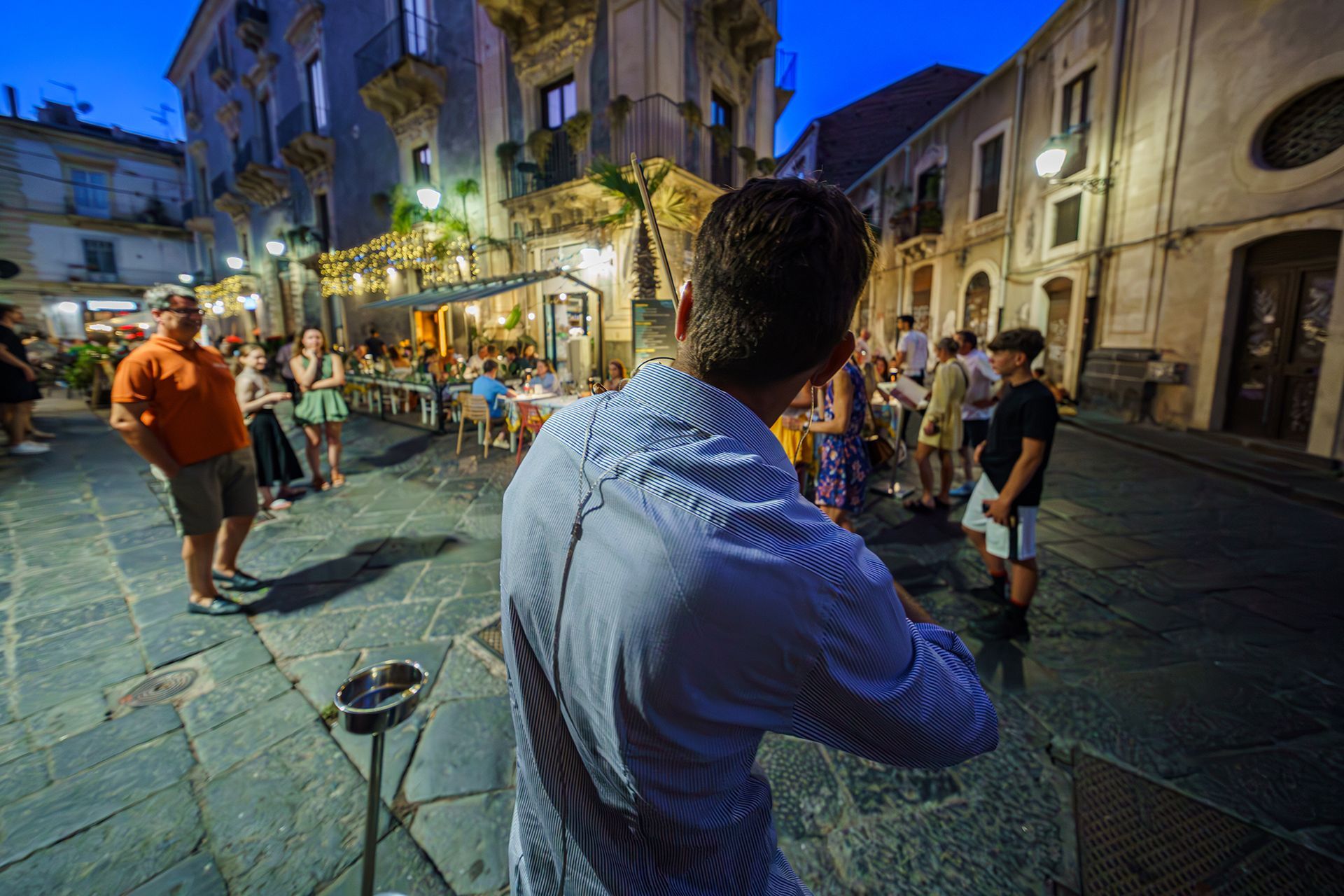 Persona con camicia blu che osserva una vivace scena di cena all'aperto in un vicolo europeo al tramonto.