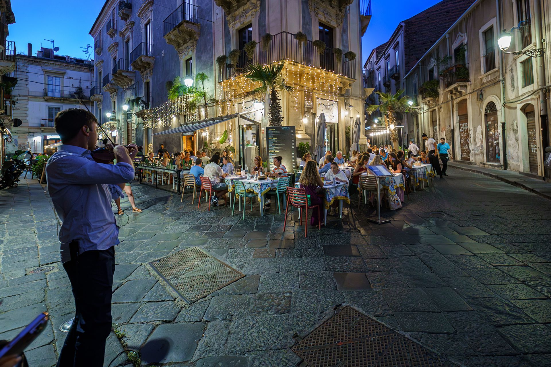 Uomo che suona il violino su una strada acciottolata di notte; persone che cenano in un bar all'aperto.