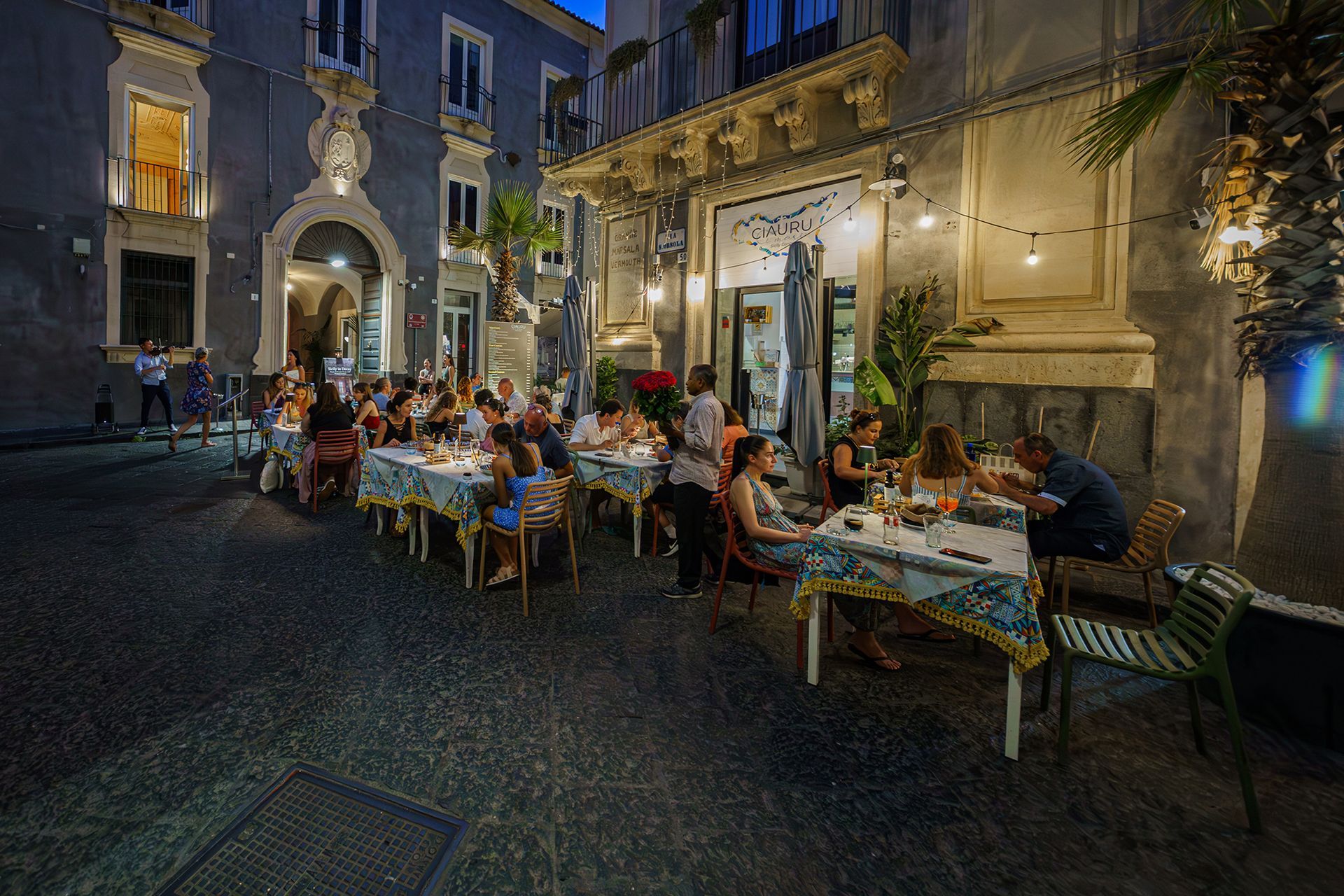Ristorante all'aperto di sera, con clienti seduti ai tavoli. Tovaglie blu e bianche. Illuminato da luci soffuse.