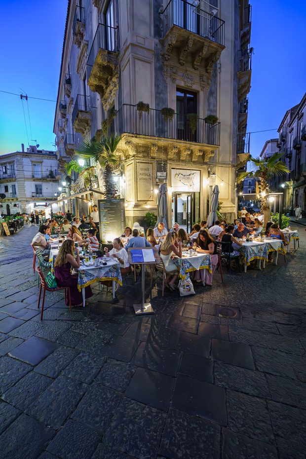 Ristorante all'aperto in una città italiana, persone che cenano ai tavoli, edificio sullo sfondo. Serata illuminata dai lampioni.