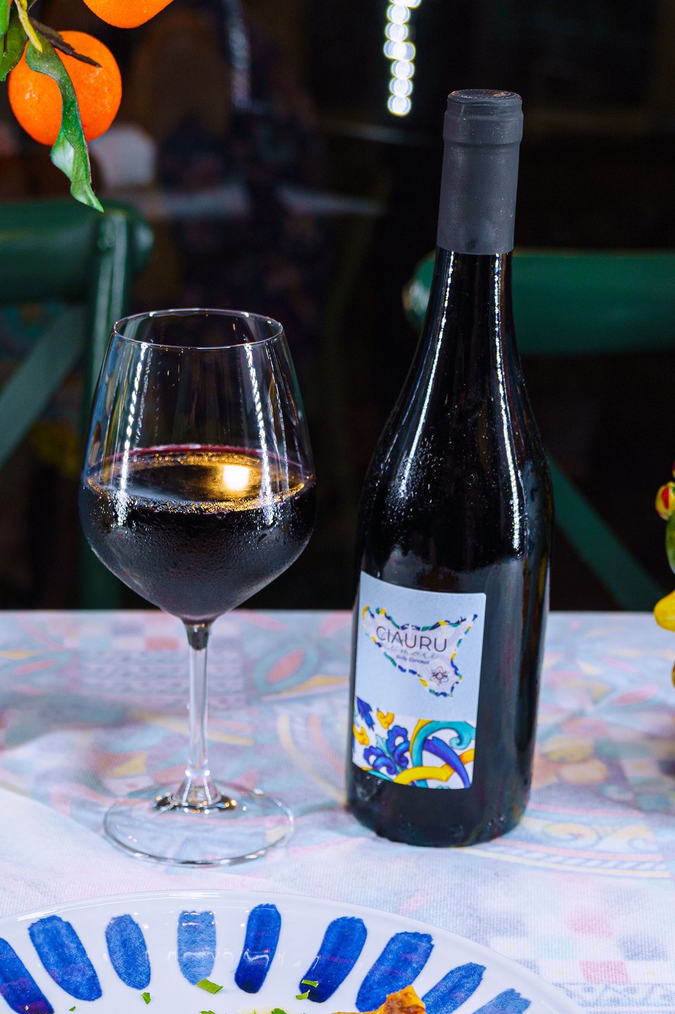 Bottiglia di vino e bicchiere su un tavolo, con un'etichetta colorata. Arance sullo sfondo.