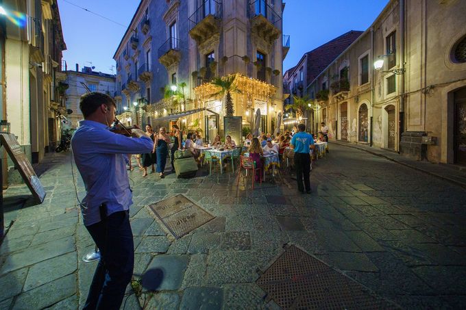Un uomo suona il violino su una strada acciottolata, i clienti del ristorante cenano al tramonto.